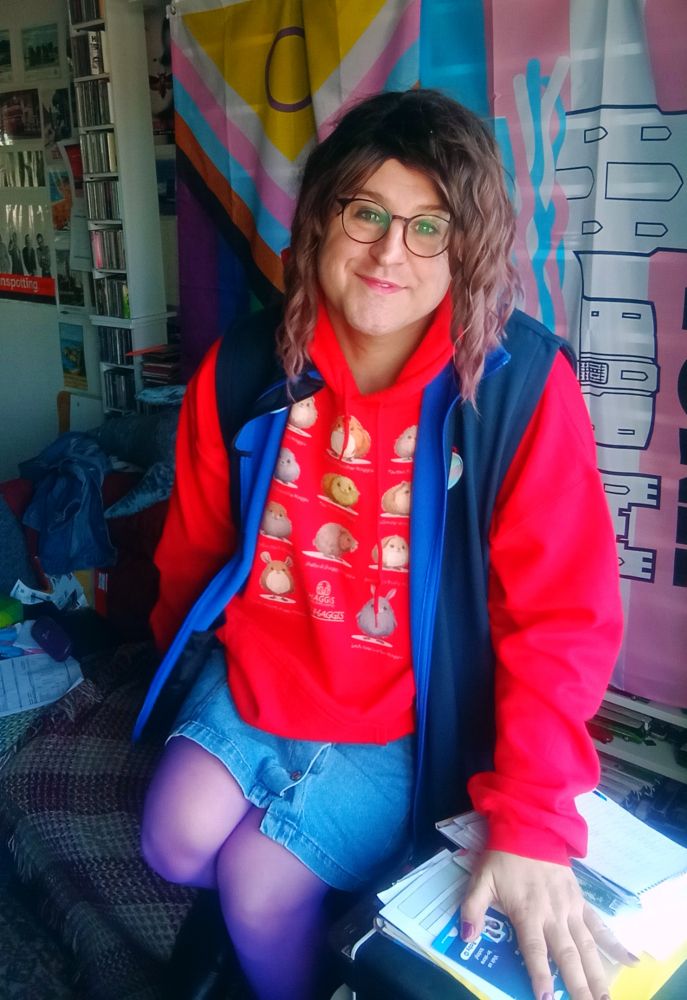 Pretty trans woman sitting on edge of sofa looking at camera. She has a red hoodie with wee yins on the front plus blue short denim skirt and purple tights. 