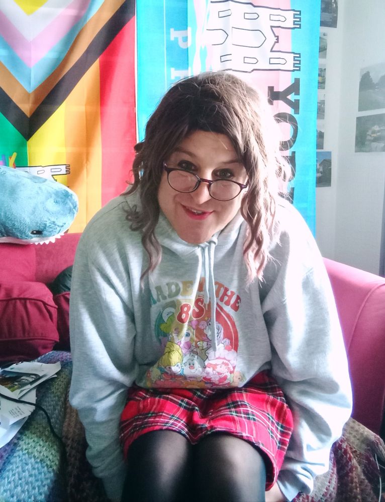 Pretty trans woman sitting on sofa in front of camera. She wears black tights,  red plaid skirt and grey hoodie. 