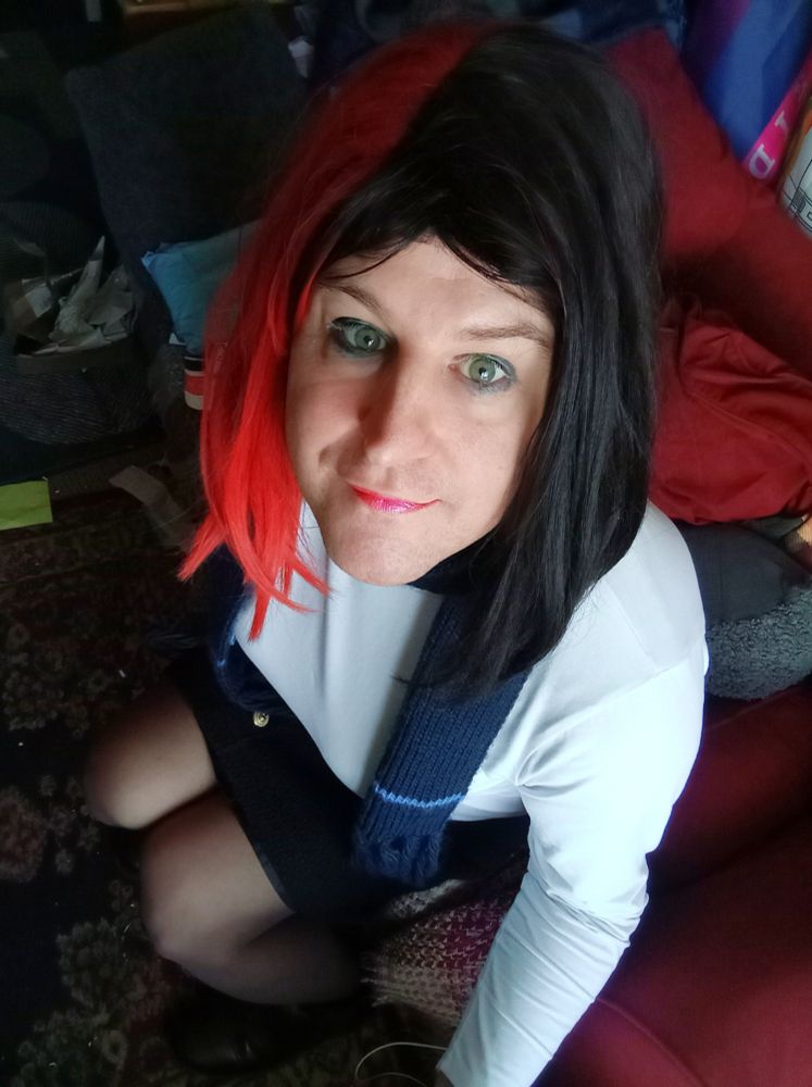 Pretty trans woman sitting looking up at camera. She has a white turtle neck top, black skirt, praline colour tights and a moderately happy facial expression. 