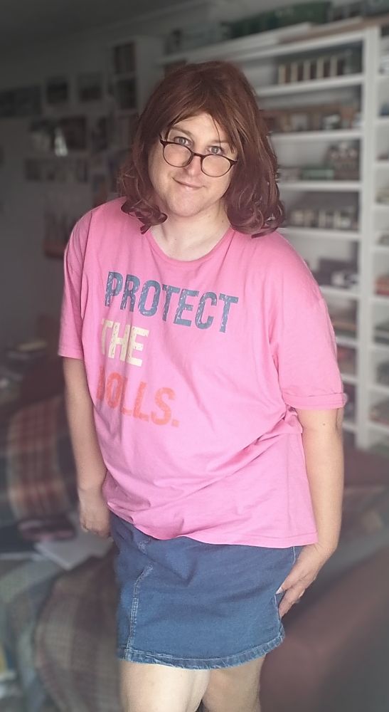 Pretty trans woman stood up. She wears a pink t shirt with 'protect the dolls' written on front and a blue denim skirt. Trans woman is also smiling. 
