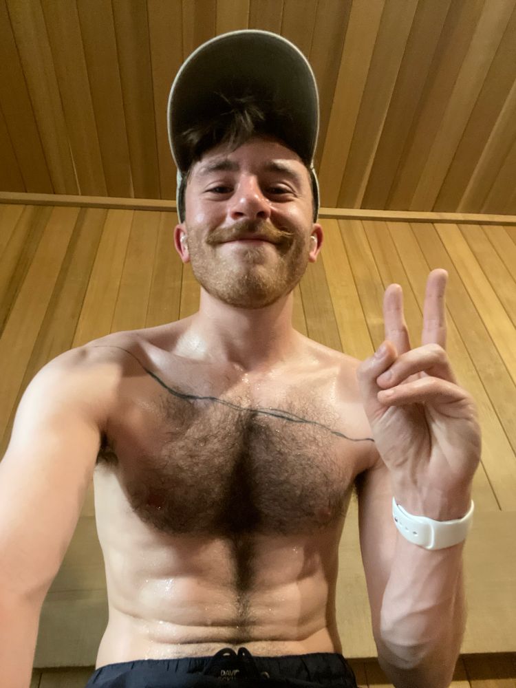 Hairy, shirtless guy with a mustache and hat holds up a peace sign while sweating it out in a sauna.