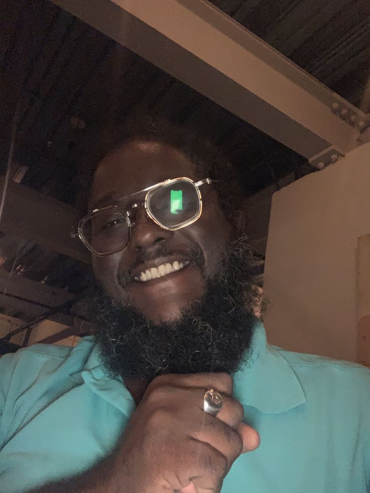 Derron Ellies, a black man from Trinidad and Tobago, smiles as he gives a right hand fist bump with a crescent & star ring on his right hand. Derron is wearing a light teal polo.