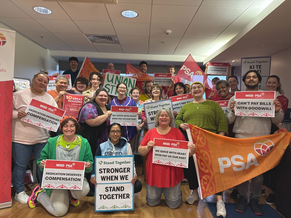  Indoors. a large group of people are facing the camera, captured mid-cheer. They are holding a variety of PSA flags, stand together and union themed placards and solidarity signs for MSA bargaining.
