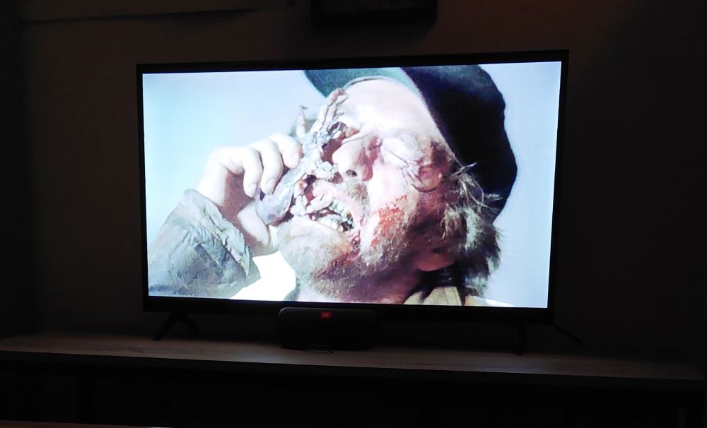 un frame du film Ticks (1993). On y voit un homme brun mal rasé portant une casquette noire. son visage a une expression de douleur et il est en train d'essayer d'empêcher une tique monstrueuse de l'attaquer