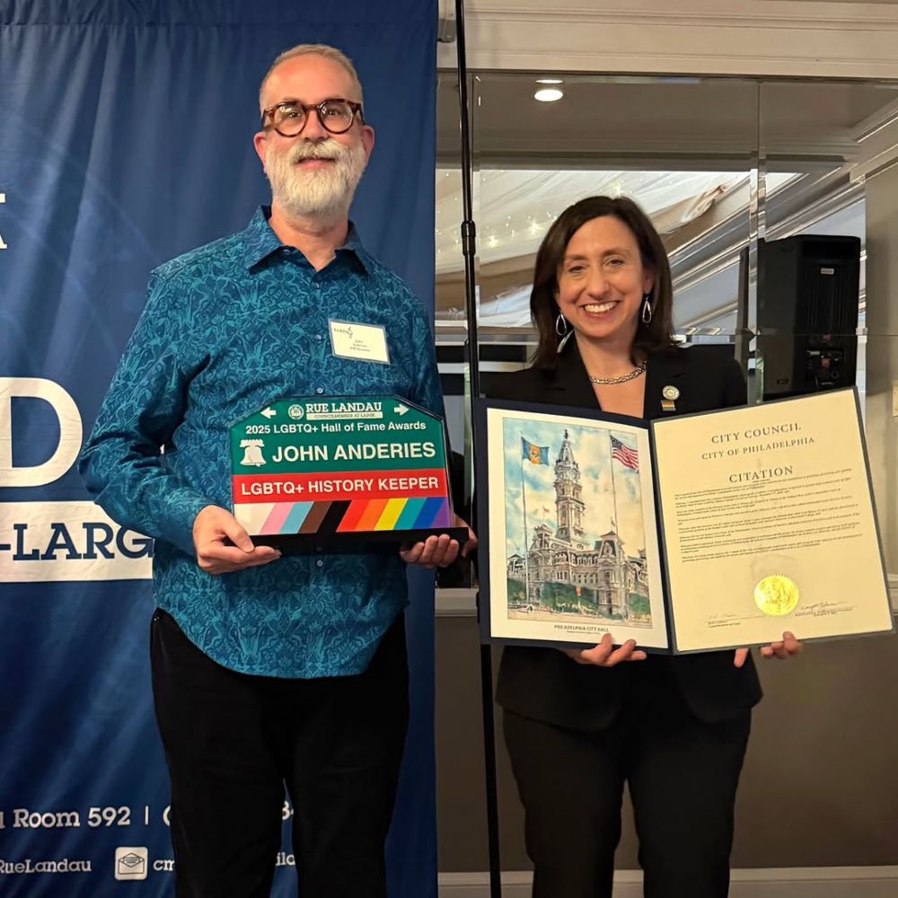 Archivist John Anderies in a turquoise shirt and dark slacks holds a rainbow street sign themed award labeled "LGBTQ+ History Keeper" while Councilperson Rue Landau in a black pants suit holds an opened citation from Philadelphia City Council.