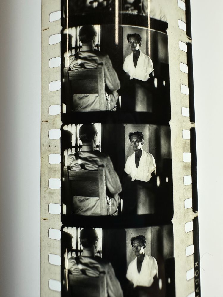 Frames from 35mm moving image film shot in black & white. Shot of two men sitting at a table in a dimly lit room. Man in uniform has his back to camera. Man in white shirt faces the camera.