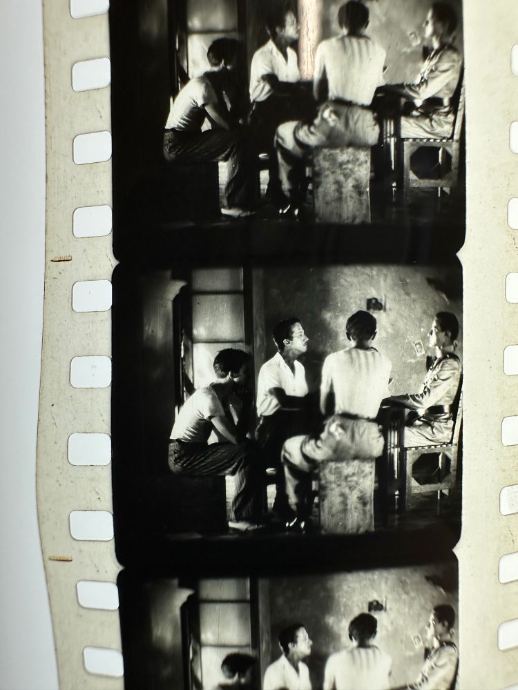 Frames from 35mm moving image film shot in black & white. Shot shows four men sitting around a table in dim light. Three men wearing white shirts. One wearing a military uniform.