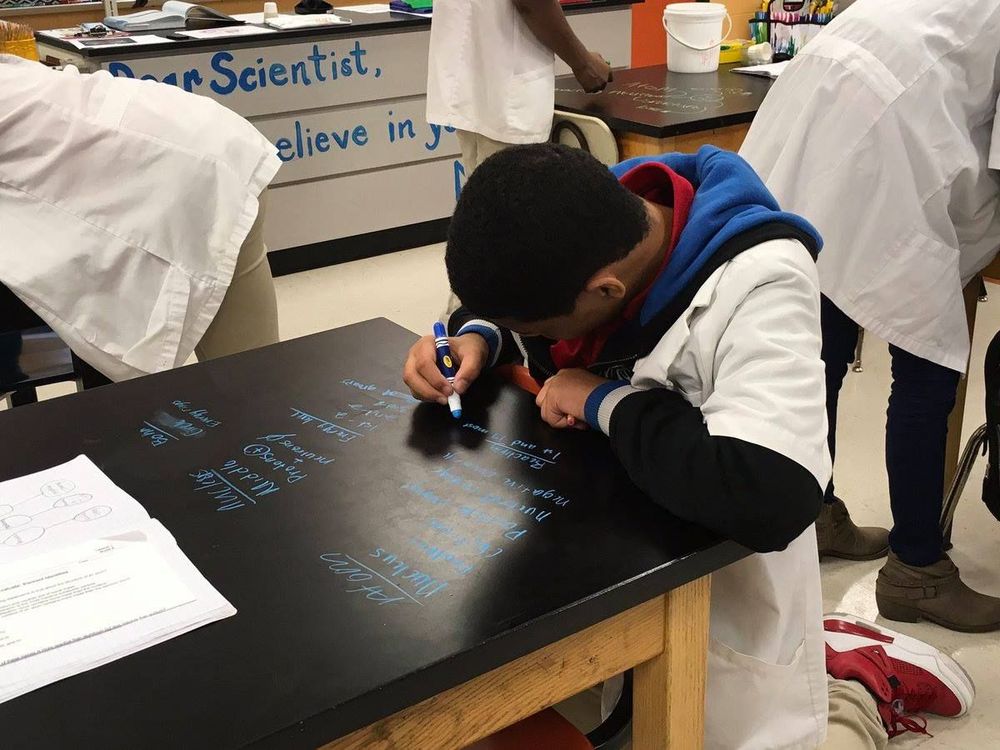 A boy in a white lab coat is writing directly on a table; his face is pointed away from the camera.