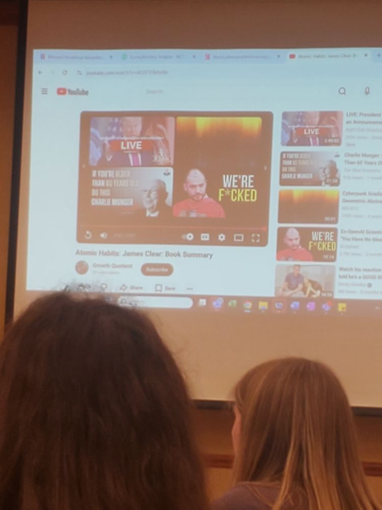 A photograph of a YouTube video on a projector screen. The video has ended and one of the recommended videos has the words "We're F*cked" in the thumbnail. The setting is a professional work conference. 