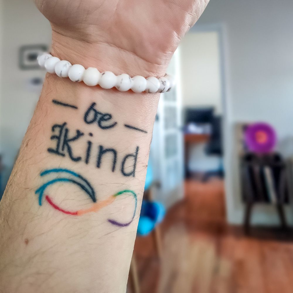 A photo of my most recent tattoo, the seventh. Located on the inside of my left wrist, it reads "—be—Kind" above a rainbow-colored Infinity symbol, one side being a cresting wave, curling towards & to complete the line's cross stroke.

"be" is set in Inkwell Script. 
"K" is set in Inkwell Blackletter. 
"ind" is set in Inkwell Sans. 