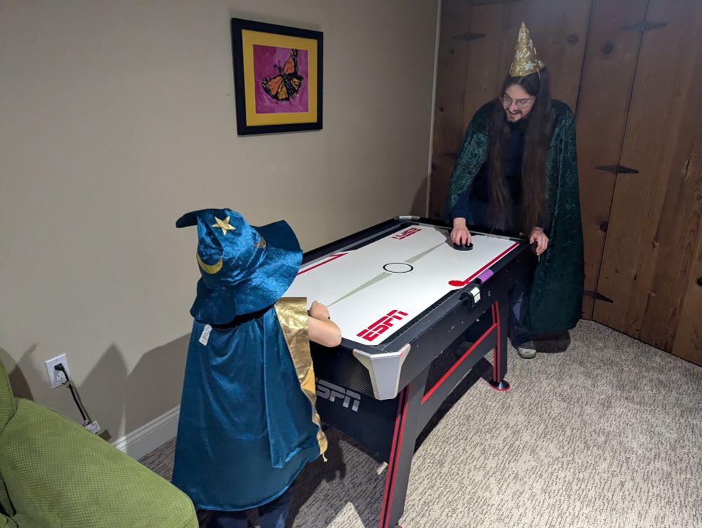 A child and adult both dressed as wizards, with wizard caps and capes, playing air hockey together.