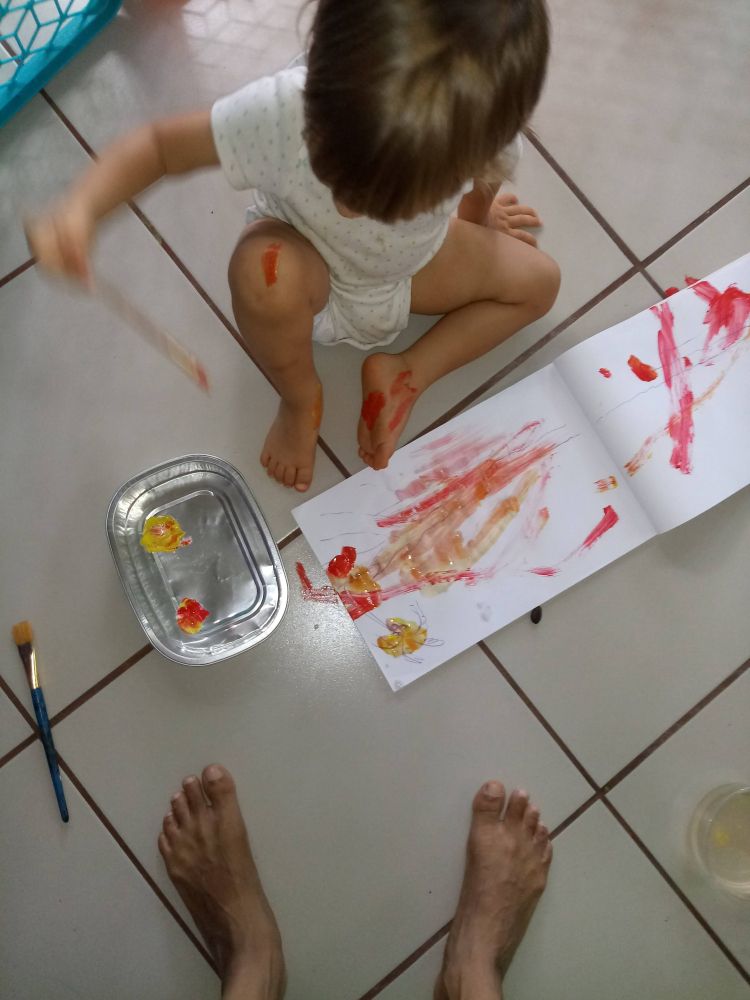 A toddler (my kid four years ago) photographed from above sitting on a tile floor with red and yellow paint in a tray and a sketchbook with red, yellow, and murky orange dashes of paint running across the pages. The sole of the toddler's left foot is painted red. There's a line of red paint on the toddler's right knee. The toddler's right hand is holding a brush and motion-blurred. More body painting will ensue.  