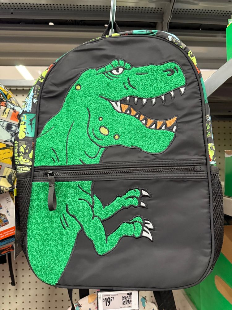 A black student backpack has a green Tyrannosaurus rex embroidered on the front. The embroidery makes it fuzzy.
