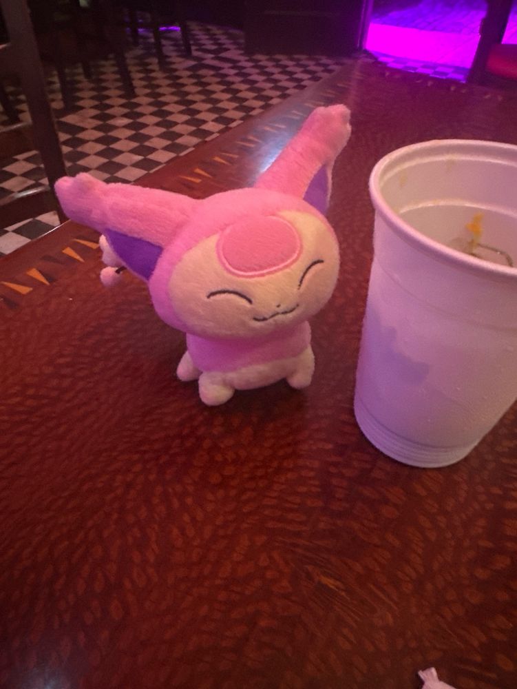skitty sits next to a plastic cup on a table. there is a checkered floor and pink lights in the background 