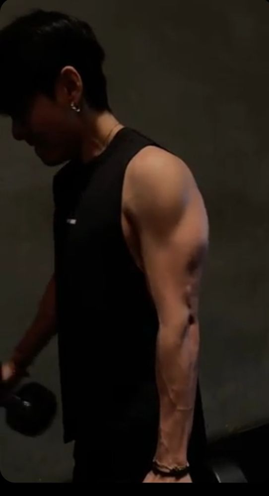 V of BTS, from a weverse moment video, in a black tank top with a gold hoop earring, overdoing it with some curls. The triceps are popping.