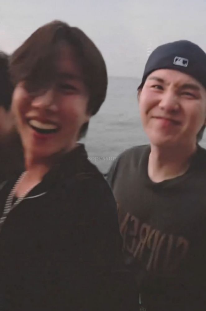 J-HOPE and SUGA of BTS from Hobi's post dinner beach patio video. Hobi has a big happy open mouth sunshinr grin, and Yoongi is behind him with a happy scrunched tsundere face.