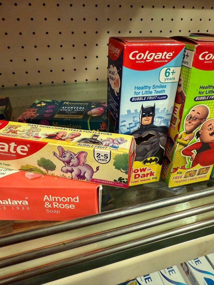 A display of various children's toothpaste and soap on a store shelf. Notable items include Colgate toothpaste for kids, featuring Batman and bubble fruit flavor, written „Glows in the dark“.