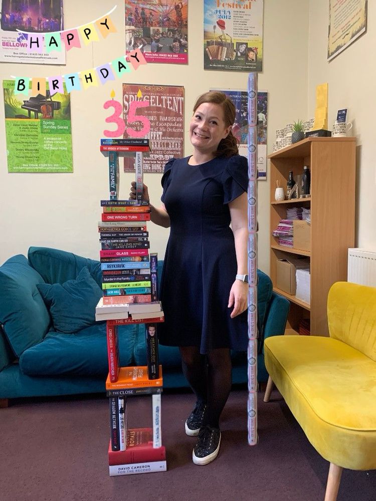 The Harrogate festival head standing next to a stack of books, which has been badly photoshopped to add a stack of one book beside her, and added candles to the top. 