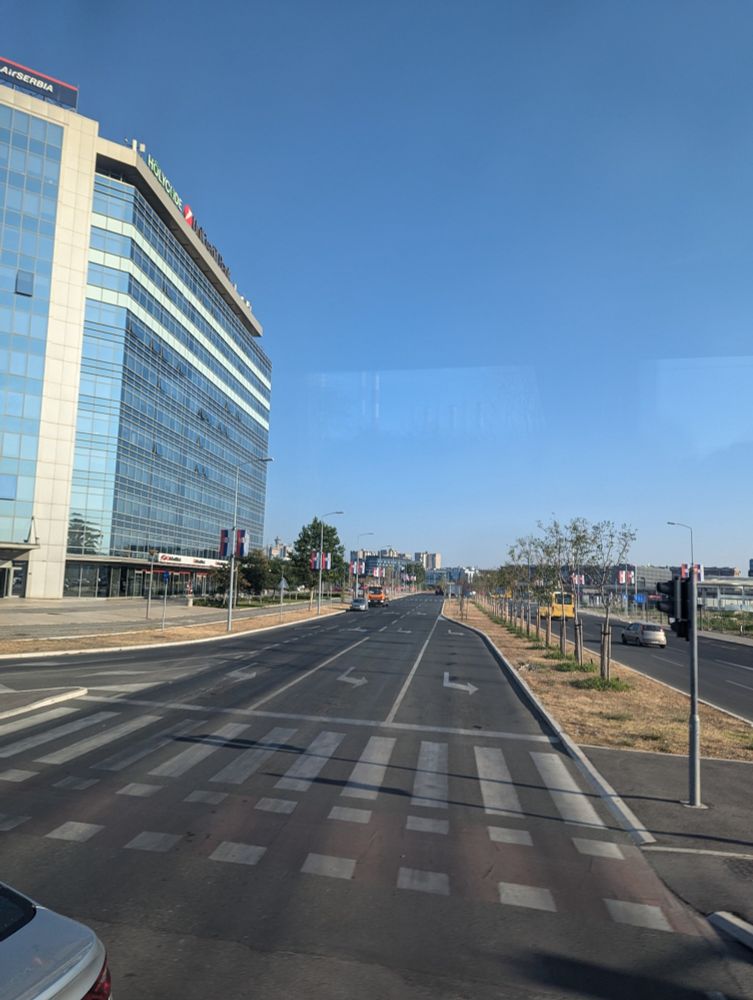 Auf den ersten Blick ein Foto einer tristen mehrspurigen Straße und daneben ein großes, verglastes Bürogebäude mit mindestens 10 Stockwerken. Im Hintergrund, recht weit entfernt, ist der Genex Tower zu sehen, und wenn man gute Augen hat oder reinzoomt erkennt man auch die ganzen Serbien-Flaggen an den Straßenlaternen.