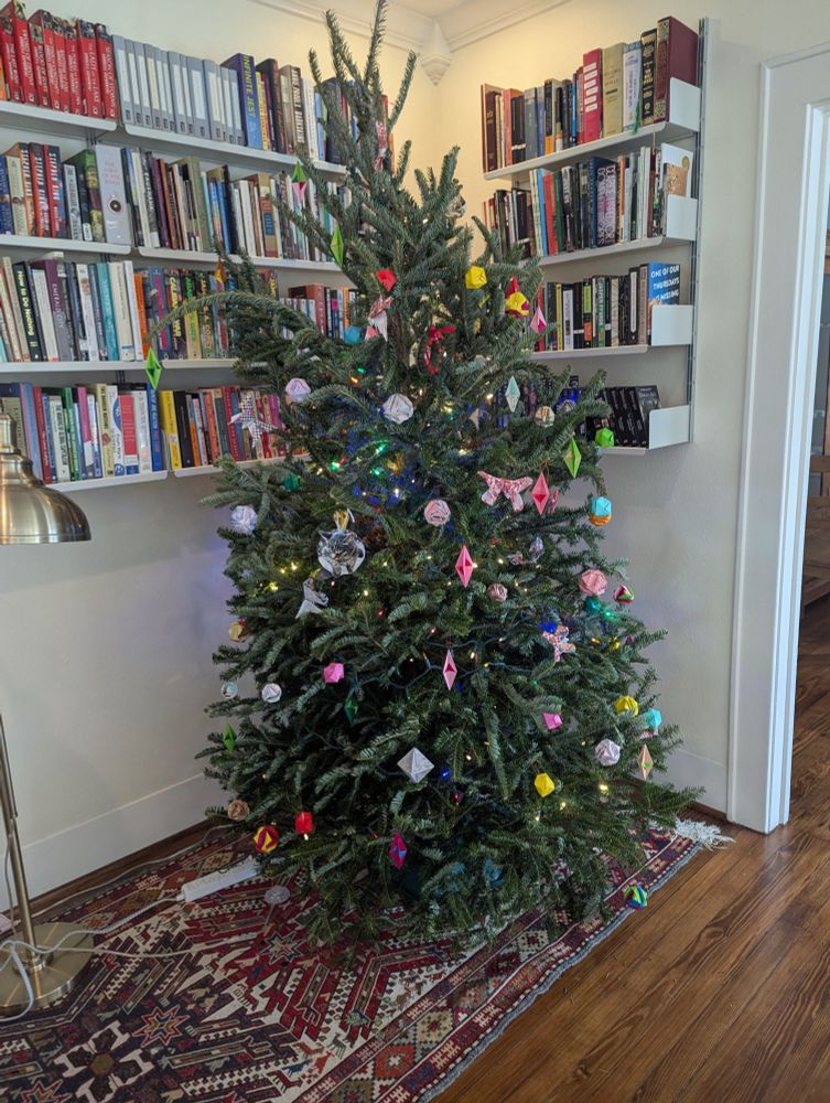 A Christmas tree covered in colorful lights  and colorful folded orgami ornaments (patterned paper and plain paper in pinks, torquoise, and other traditional Christmas colors). Ornaments include LaFosse butterflies, diamond pendant shapes, water balloons, and brocade balls. 