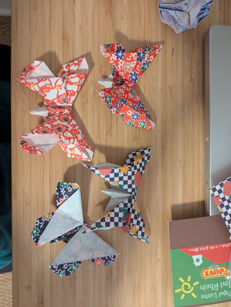 Four different origami butterflies (Michael LaFosse models) in patterned paper: red with blue and golden flowers, white with red and white flowers with navy center, red and yellow flowers with a checkerboard background, and blue with green and gold flowers. 