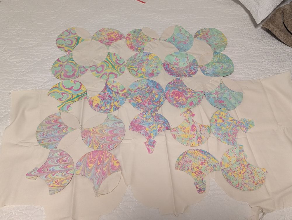 Partially handpieces quilt top made of clamshells and points. Some of the pieces are  hand marbled in pink, blue, yellow, and green, some are off white. Half the marbled pieces haven't been sewn together and are simply placed on the off white fabric backdrop. 