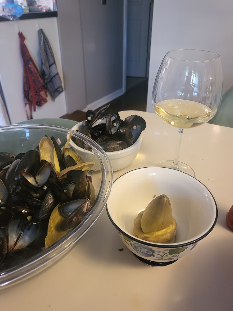 The view of a table with a bowl of muscles shells and a small bowl with an artichoke heart. A glass of white wine is in the background