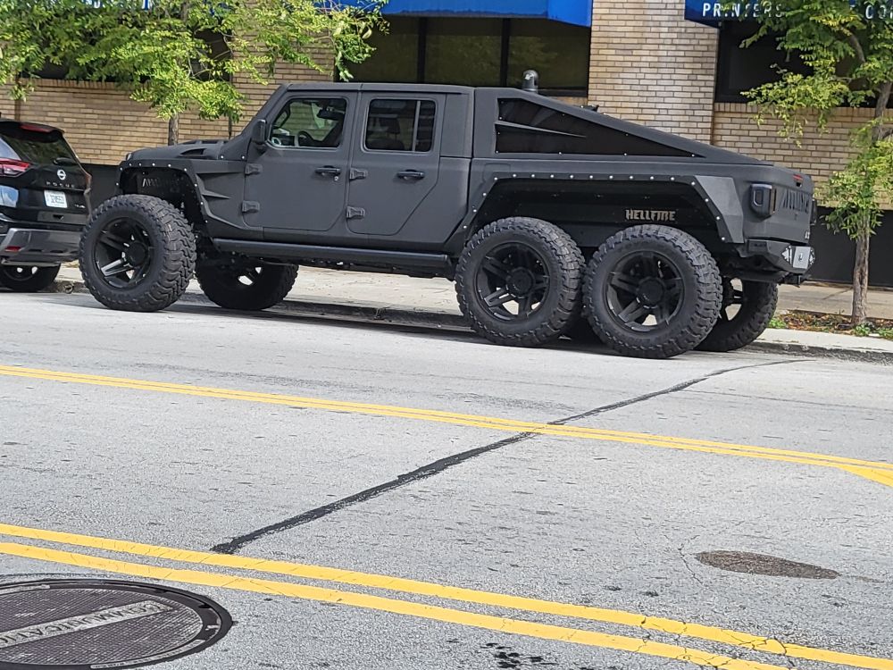A six wheeled jeep with giant wheels and a long pickup bed. The word, hellfire is visible in the rear wheel well. Parked in Printers Row, downtown Chicago. 