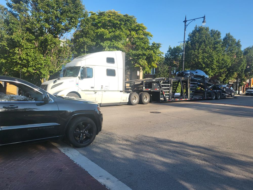 White semi tractor with a carcarrying trailer. A black sedan is on the upper level of trailer and a white compact car is being loaded on behind it. The semi-trailer are blocking an crosswalk