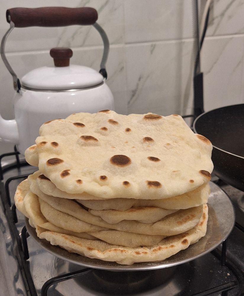 Uma pilha de pães sírios sobre um prato inox, sobre o fogão, uma chaleira branca ao fundo.