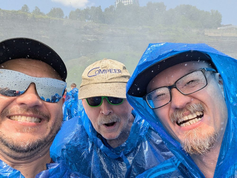 Three men in blue plastic ponchos smiling in mist near Niagara Falls