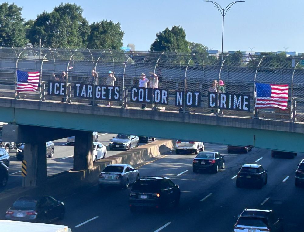 People displaying banner over a bridge reading "ICE targets color not crime" and an American flag on either side.
