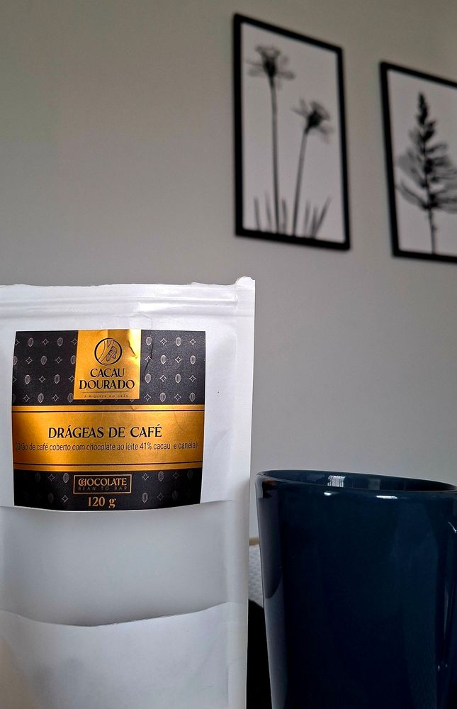 Embalagem branca com rótulo dourado escrito "Drágeas de café" da marca Cacau Dourado. Ao lado, uma caneca cinza escura. Ao fundo, parede branca e quadros de plantas preto e branco.
