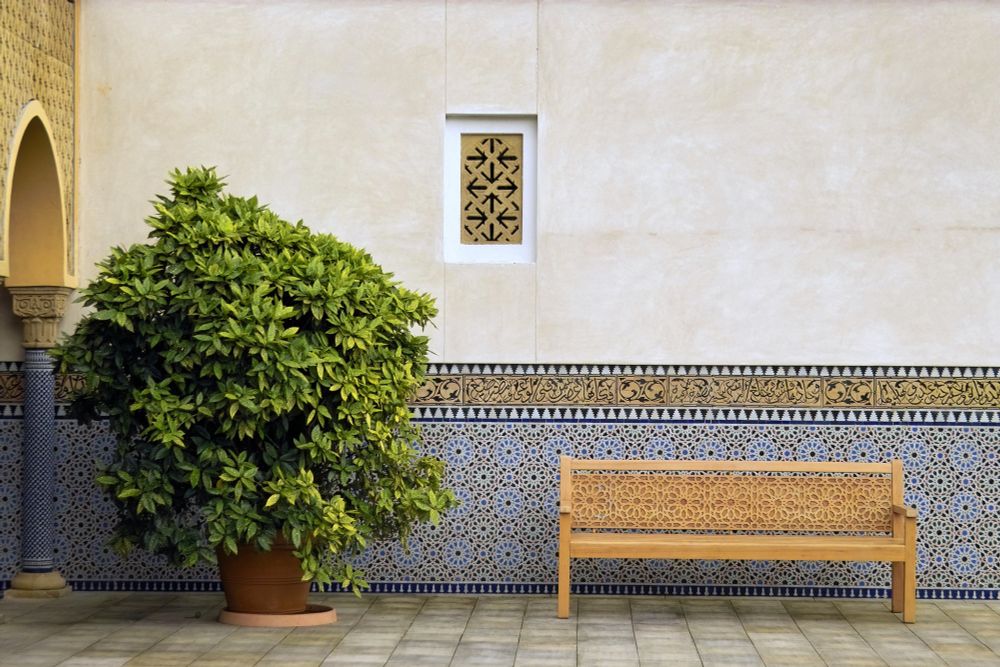 Blick auf eine mit Kacheln verzierte Wand im Orientalischen Garten. Davor steht eine Bank aus Holz mit ornamentalen Verzierungen. Auch das kleine Fenster ist verziert mit einem Muster. Links steht ein grüner Busch, Zitronenbusch.  