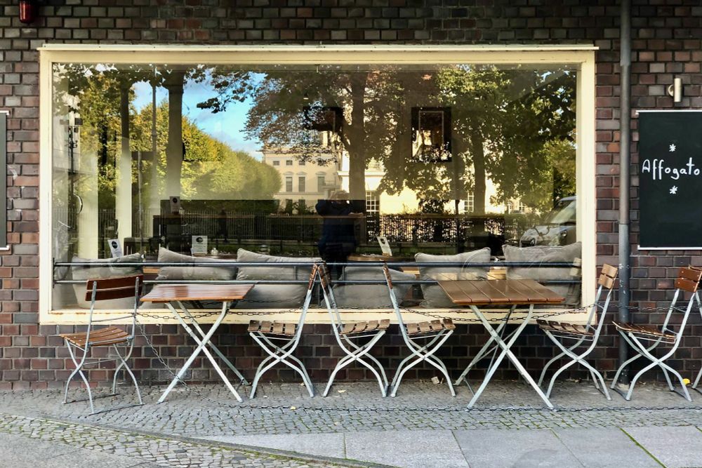 fronaler Blick auf ein Gebäude mit Backsteinfassade und einem sehr großen und breiten Fenster. Davor stehen mehrere filigrane Stühle und Tischedas Metall mit Holzteilen. Das grpoße Fenster spiegelt den gegenüber liegenden Park mit Bäumen und einen Teil des Charlottenburger Schlosses.
