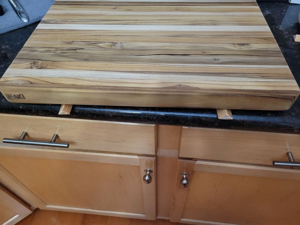 A picture of two triangular slats of wood perfectly supporting a teak cutting board on a counter, so it can be oiled and left to dry