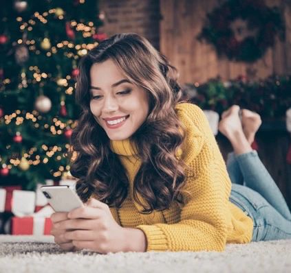 a woman, beautiful & bemused, looks at her phone while lounging at the foot of a tastefully decorated christmas tree