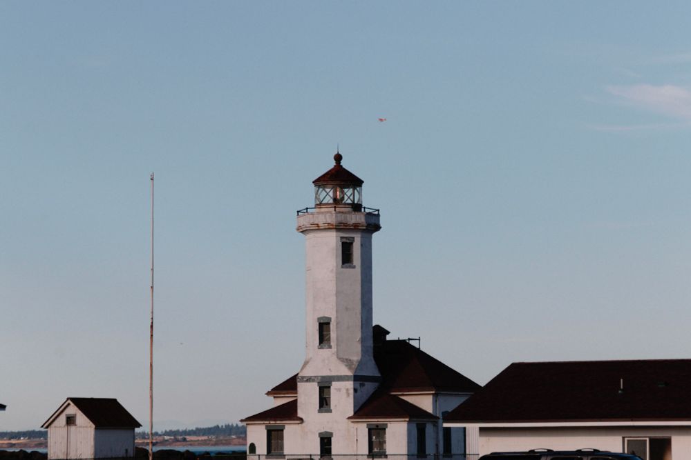A tall, white lighthouse with a red roof under a clear blue sky. Nearby there are smaller buildings with a similar roof.