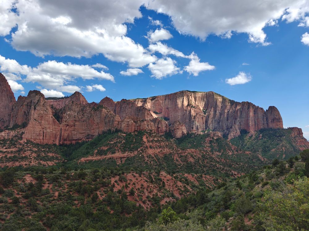 A mountain range with blue sky, white clouds, and green trees 