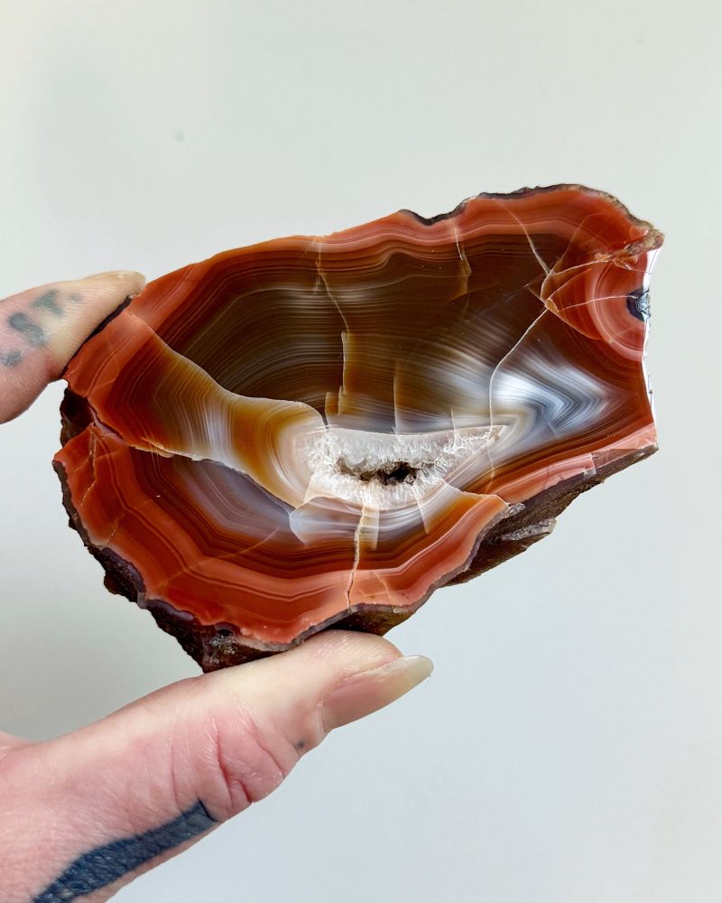Photo of a two finger holding a polished agate half against a plain white background. The agate displays angular concentric (fortification) banding around the outer edges, and banding ranges in colour through red, brown, grey, white, orange and pale blue. There’s also an eye on the right hand edge where there’s banding forming in a concentric circle. The centre exhibits a small vug with white to clear/colourless crystalline quartz and small quartz crystals which point inwards. From south of Arbroath, Angus. Held in the Hunterian Collections, University of Glasgow.