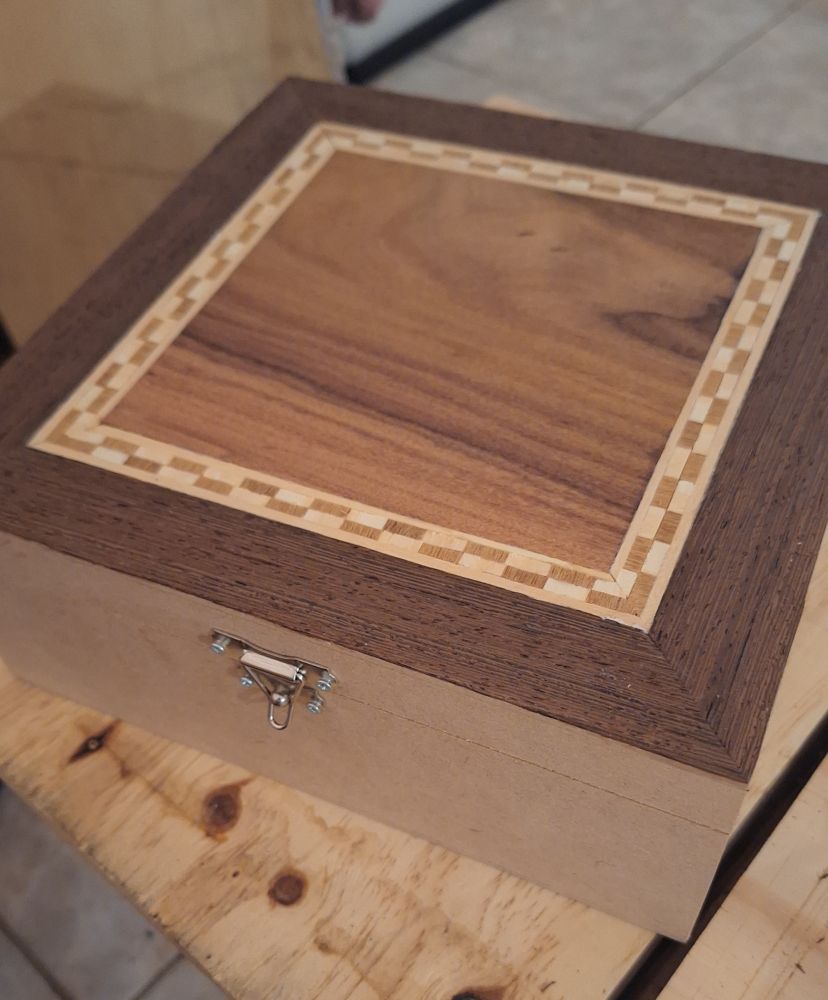 Caixa quadrada de MDF com marchetaria feita na parte de cima da tampa. A marchetaria é um quadrado com veios da madeira bastante visíveis, um pouco menor que a caixa, emoldurada por um padrão quadriculado/listrado e há mais uma moldura num tom mais escuro completando o tamanho total da caixa