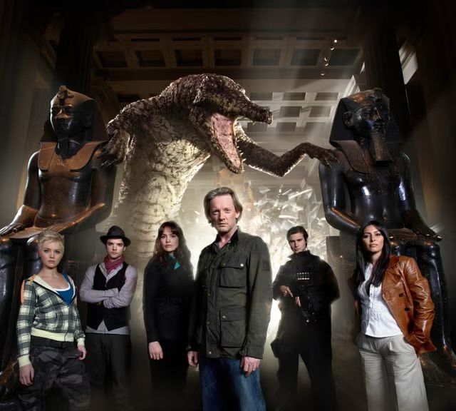Six people stood in front of a CG dinosaur. All are dressed in very standard, non fancy street clothes 
