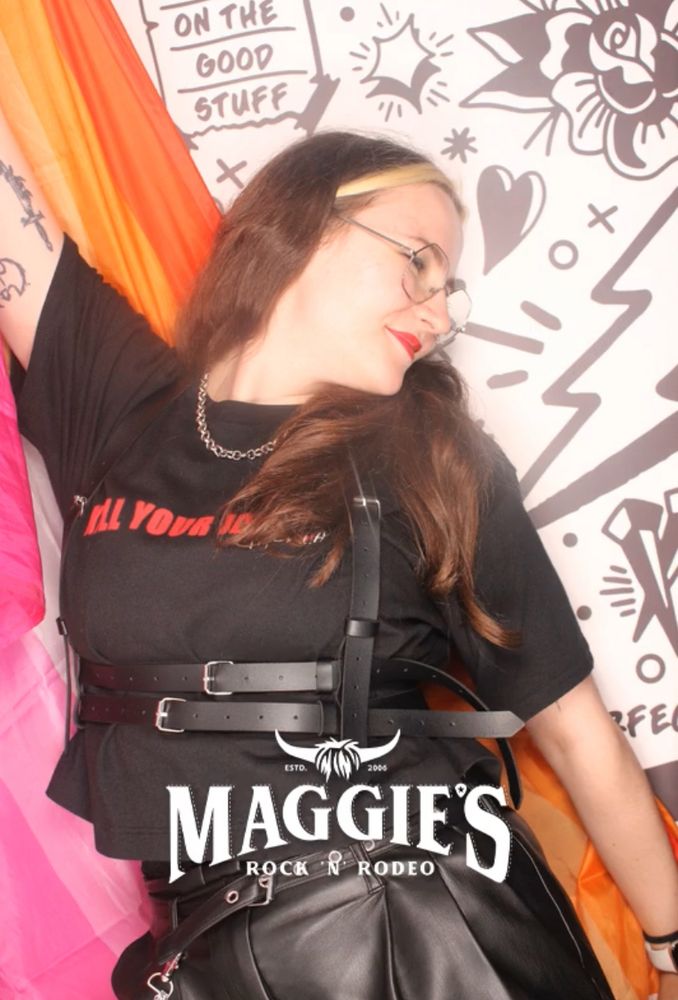 bex in the same outfit in a photo booth - they have a lesbian flag around their shoulders and are looking down to the right with a small smile 