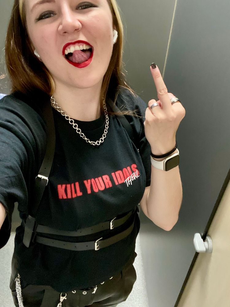 bex in a black shirt reading “kill your idols” along with leather shorts and a black harness. they’re flipping off the camera and have dark eyeliner and red lipstick on. 