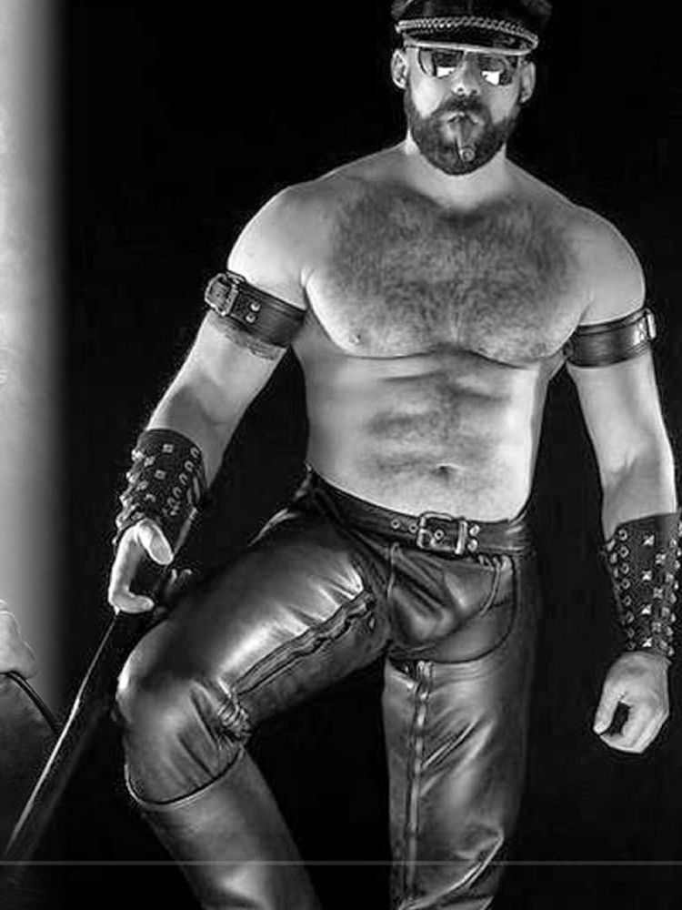 Shirtless man who may be classified as a "bear" in BDSM wear. Leather pants and leather cap wearing sunglasses and smoking a cigar. Also wearing leather vambraces and bicep straps. 