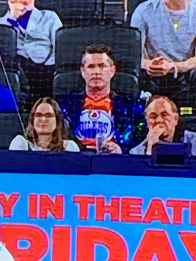 A man is seated, wearing a blue sequined blazer with an orange Edmonton Oilers t-shirt underneath. He has short dark brown hair and is holding a beverage in his left hand 