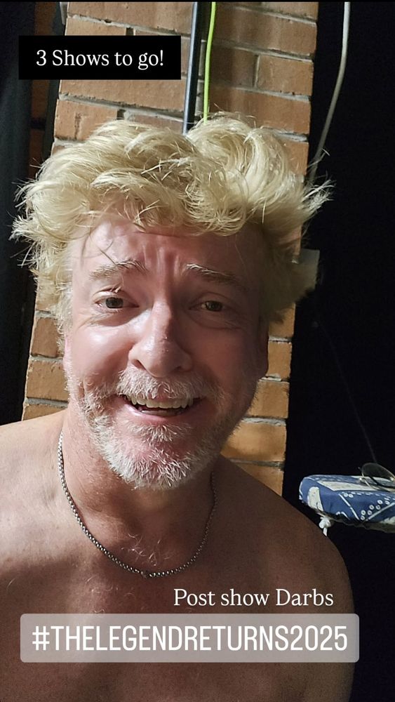 Shirtless Rhys Darby smiling at the camera with ruffled up hair and a slutty little chain and the caption post show darbs 3 shows to go