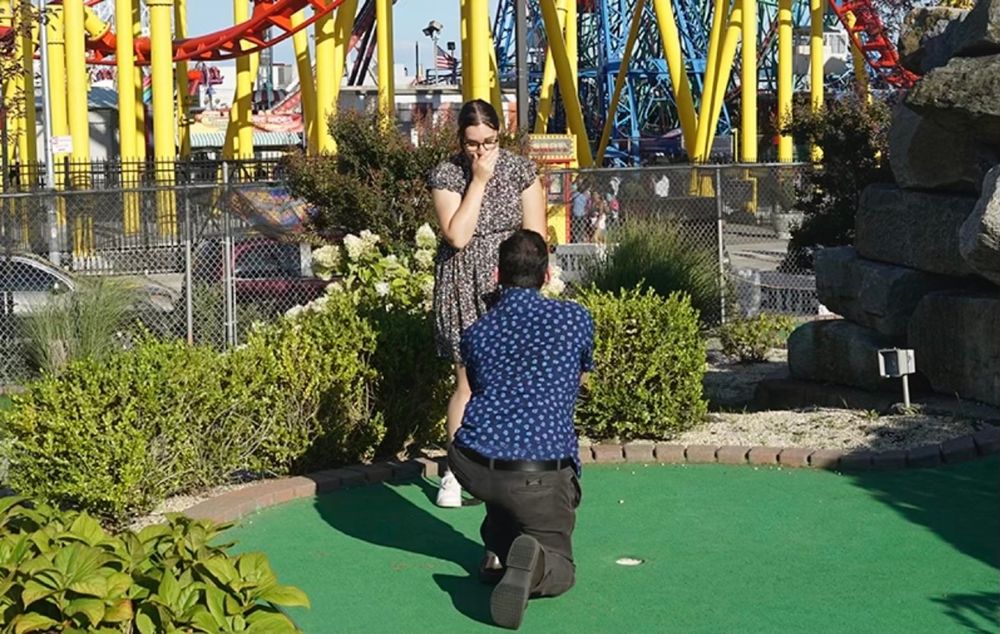 A man in blue shirt bending down on one knee in front of a woman in a floral dress, on a miniature golf course at Coney Island. A roller coaster can be seen in the background.