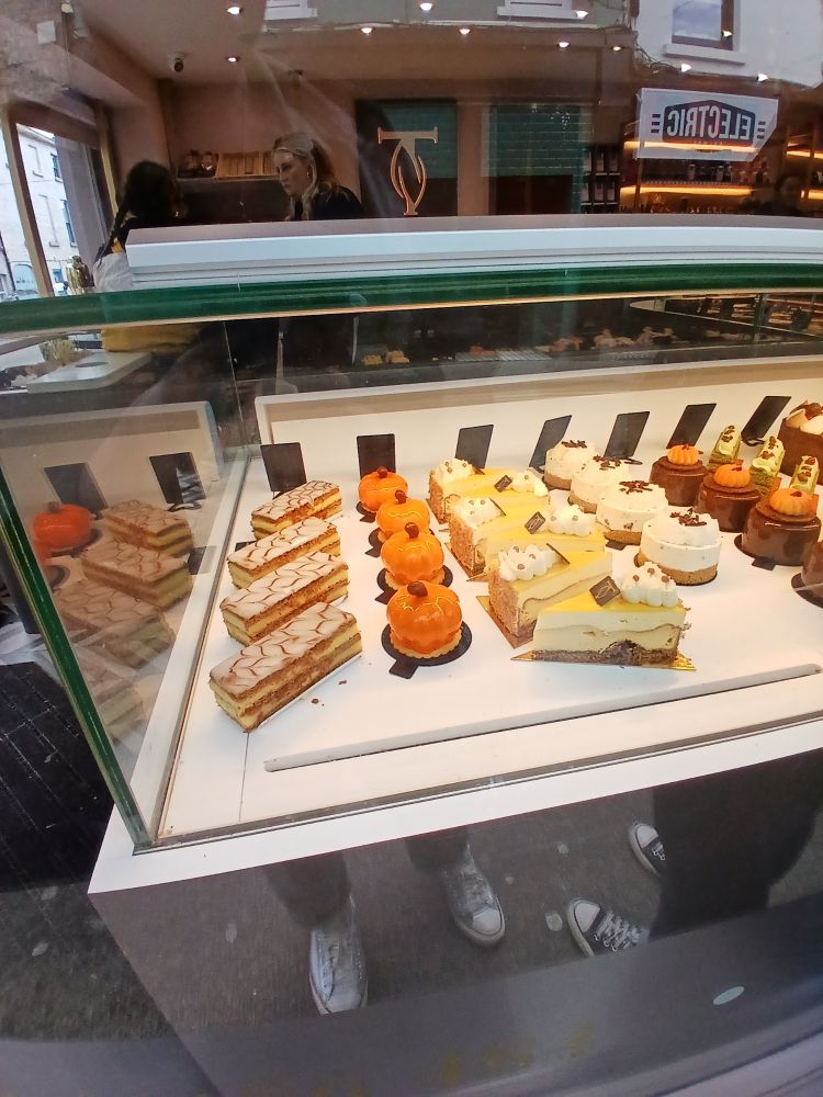 A selection of delicious patisseries in a shop window, including custard slices, individual cheesecakes, pumpkin shaped cakes, glossy cakes etc. You can see my feet reflected in the window.
