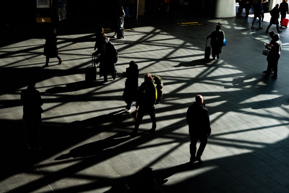 Low-light, high-contrast photo of travellers on station concourse casting small patches of colour onto the largely monochromatic image; geometrical dark shadows being cast across the floor from large window grid.